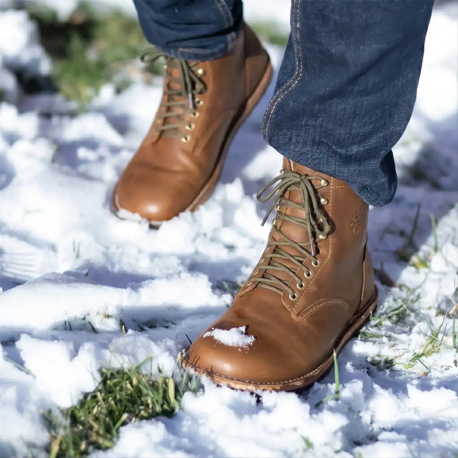 Brown leather lace-up boots with gold-toned eyelets and laces, worn by someone in blue jeans standing on snow.